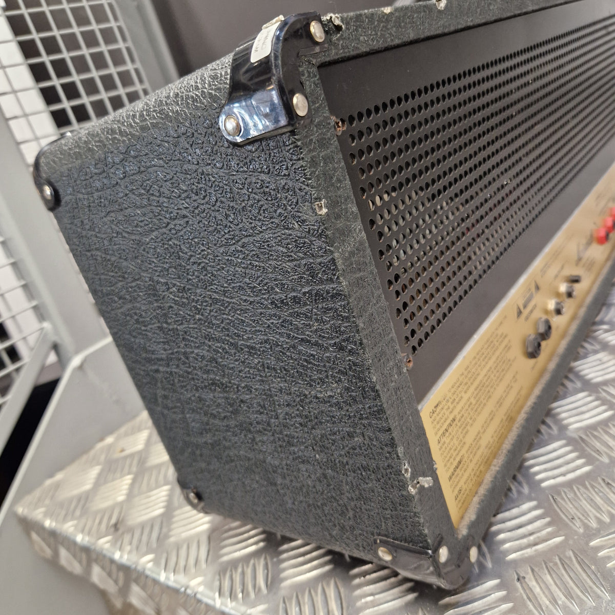 Pre-Owned Marshall JCM2000 DSL100 Valve Electric Amplifier Head A close-up of a black textured speaker cabinet corner with metal lugs and a perforated metal top grille, photographed against a diamond-plate metal background.