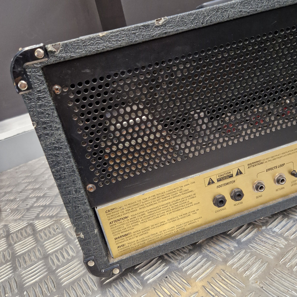 Pre-Owned Marshall JCM2000 DSL100 Valve Electric Amplifier Head Close-up of the back panel of a black guitar amplifier or speaker cabinet, showing a perforated metal grille, control knobs, and a yellow specification label on a diamond plate metal surface.