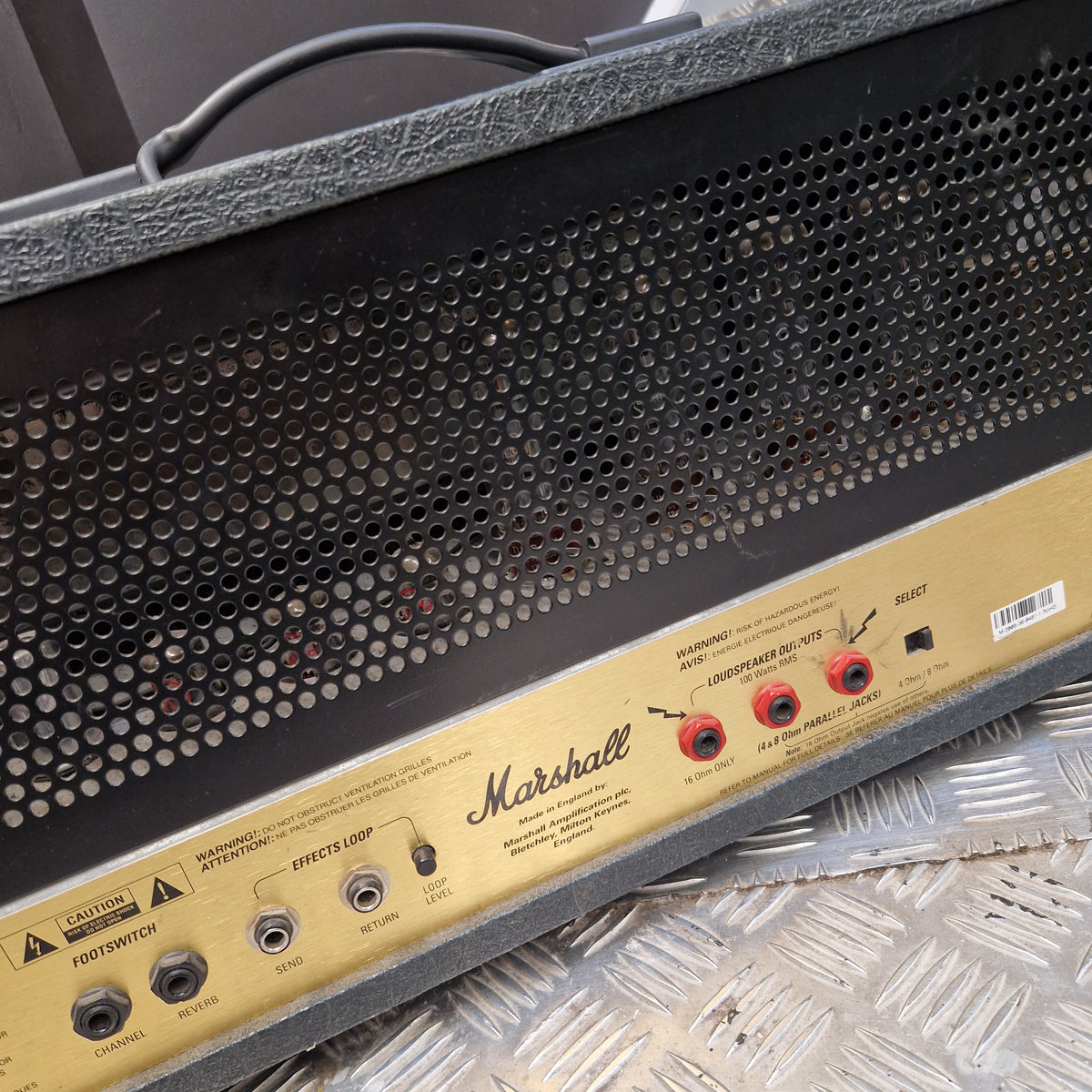 Pre-Owned Marshall JCM2000 DSL100 Valve Electric Amplifier Head Close-up of a Marshall amplifier front panel with a black perforated grille and a gold control panel showing red and black input/output jacks.