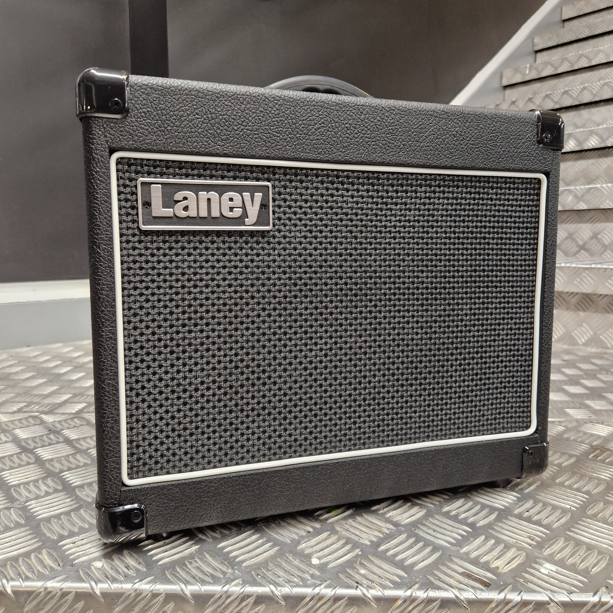 Pre-Owned Laney LG20R 20w Electric Guitar Combo A black Laney speaker cabinet with a textured front grille and a silver brand badge, photographed on a diamond plate metal surface.
