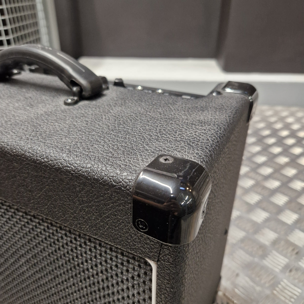 Pre-Owned Laney LG20R 20w Electric Guitar Combo Close-up of a black textured speaker cabinet corner with a glossy black protective cap and a metal mesh grille on a silver metal platform.