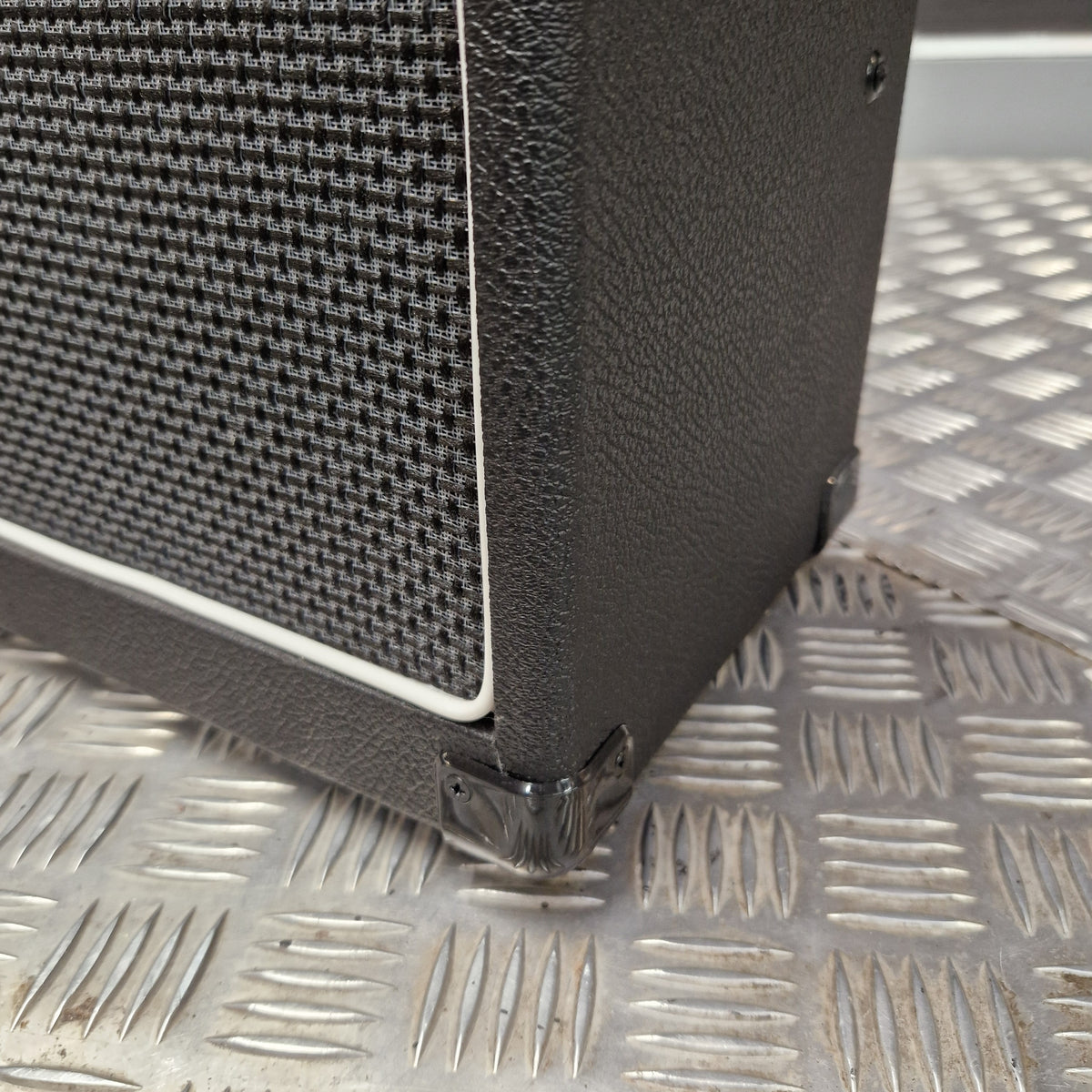 Pre-Owned Laney LG20R 20w Electric Guitar Combo Close-up of a black textured speaker cabinet corner with a black mesh grille and white edge trim, photographed against a silver diamond-plate metal background.