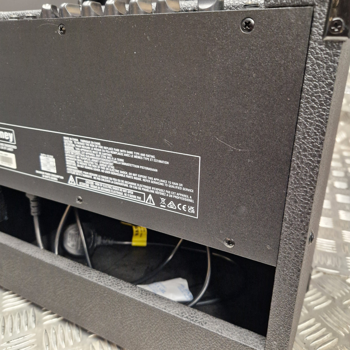Pre-Owned Laney LG20R 20w Electric Guitar Combo Close-up of the underside of a black equipment case or speaker cabinet, showing screws, a product info label, and internal wiring in a metallic diamond-plate floor setting.
