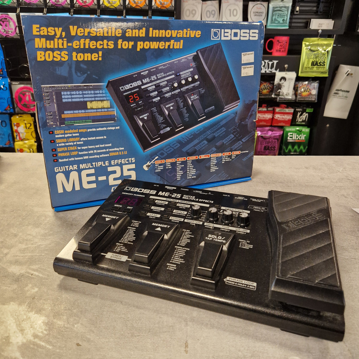Pre-Owned Boss ME-25 Multi-Effects Pedal BOSS ME-25 guitar effects pedal and its retail box displayed on a store floor, with other products visible in the background.