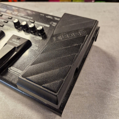 Pre-Owned Boss ME-25 Multi-Effects Pedal A close-up of a black guitar effects pedal, showing the textured footswitch and part of the control panel with knobs, resting on a gray surface.
