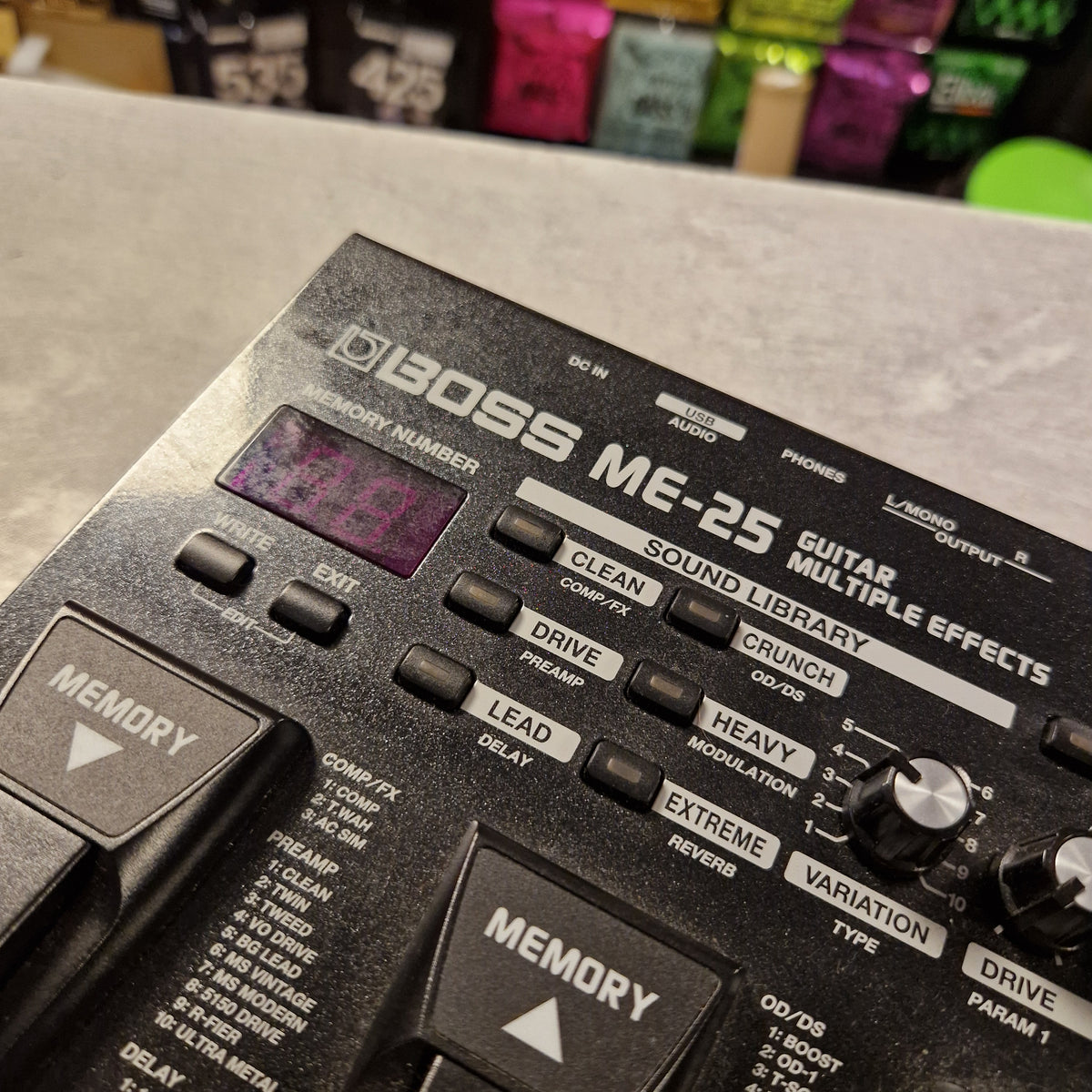 Pre-Owned Boss ME-25 Multi-Effects Pedal A close-up angled shot of a Boss ME-25 guitar modulation effects pedal, showing its black plastic body, silver and white text, knobs, and footswitch buttons. The pedal is resting on a gray surface with a blurred background containing colorful items.