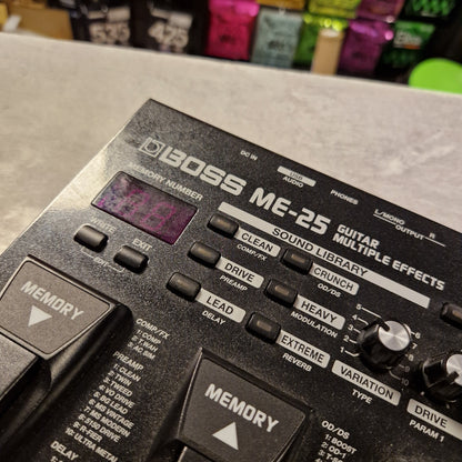 Pre-Owned Boss ME-25 Multi-Effects Pedal A close-up angled shot of a Boss ME-25 guitar modulation effects pedal, showing its black plastic body, silver and white text, knobs, and footswitch buttons. The pedal is resting on a gray surface with a blurred background containing colorful items.