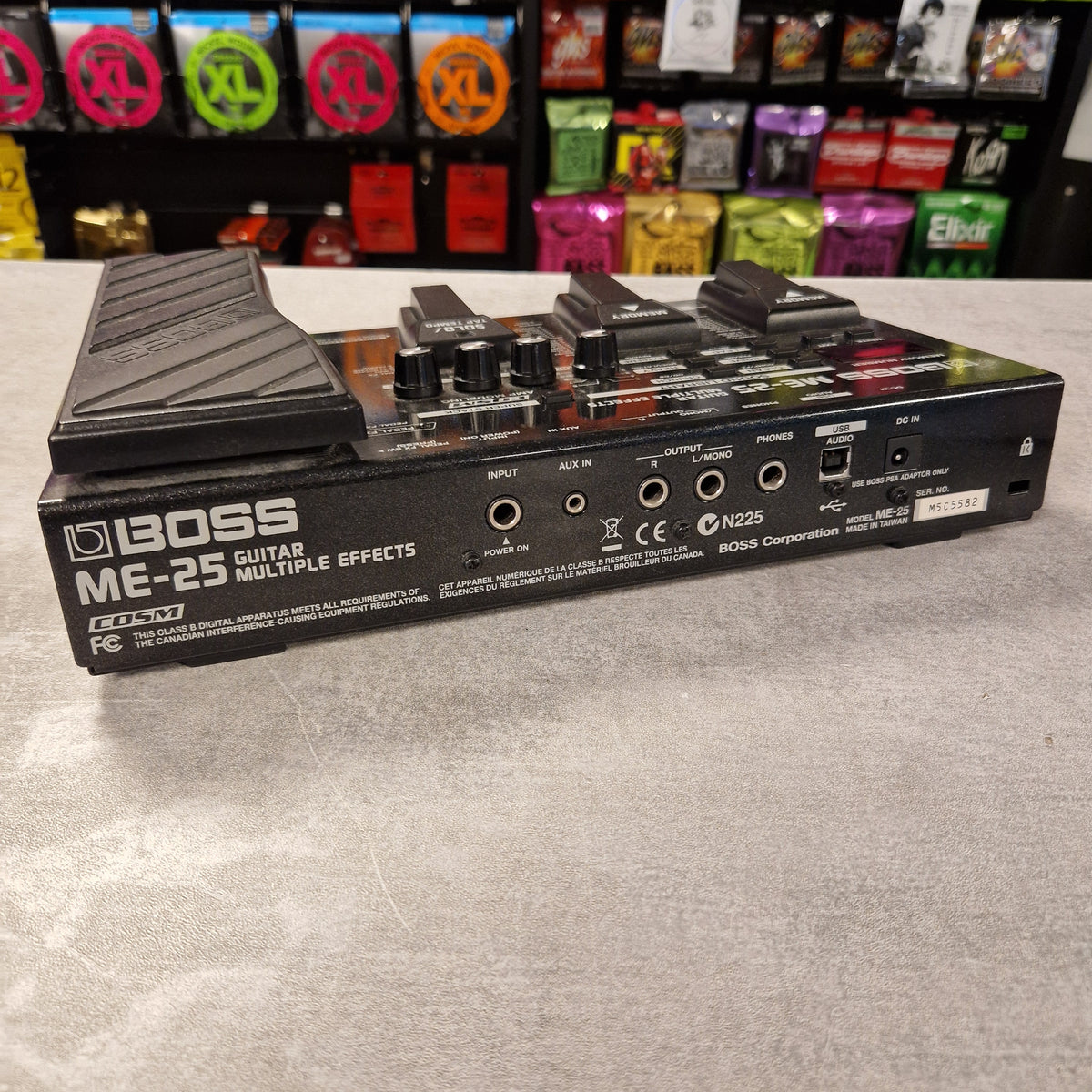 Pre-Owned Boss ME-25 Multi-Effects Pedal A Boss ME-25 Multiple Effects pedalboard shown at an angle on a light gray surface, with a store shelf of colorful items blurred in the background.