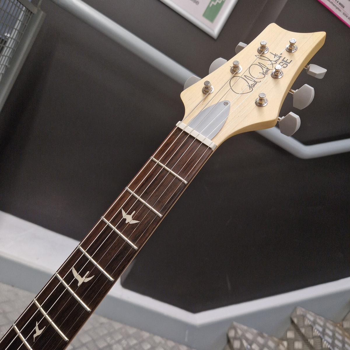 Pre-Owned PRS SE Silver Sky John Mayer Signature Electric - Dragonfruit w/ Gig Bag - Close-up of a guitar headstock and fretboard, showing a light natural wood headstock with six silver tuning pegs, a white pickguard, and a dark brown fretboard with silver bird inlays. The brand name 'KIKIKI' is printed on the headstock.