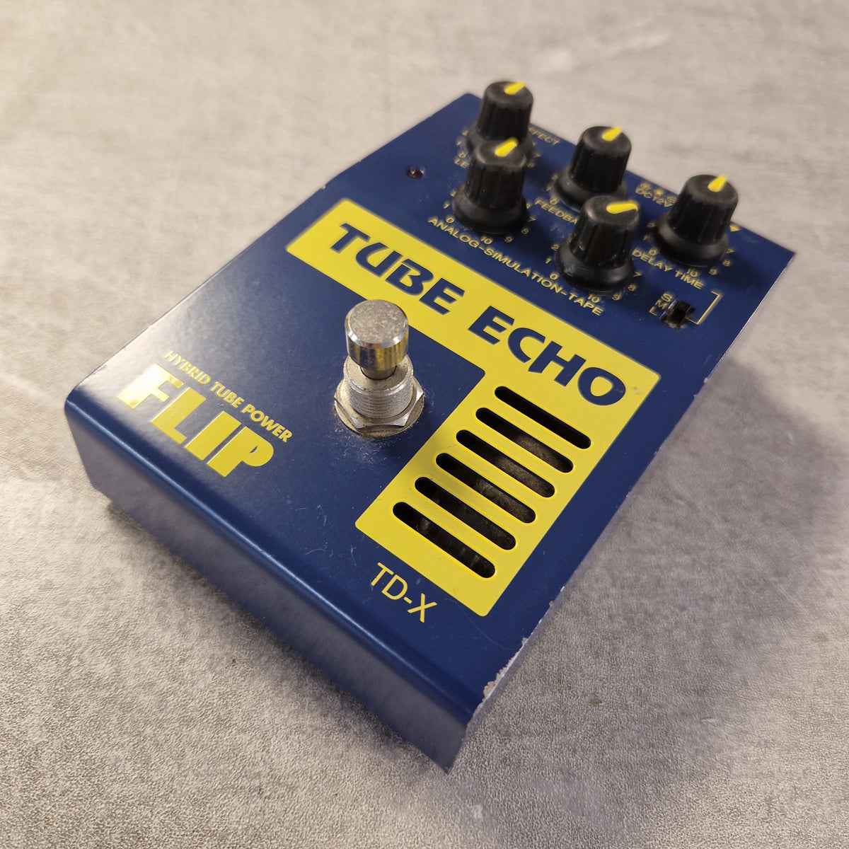 Pre-Owned Guyatone Flip TD-X Tube Echo Hybrid Tube Power Delay Pedal - A blue and yellow FLIP Tube Echo FX pedal with four black knobs and a silver footswitch, photographed on a gray textured surface.