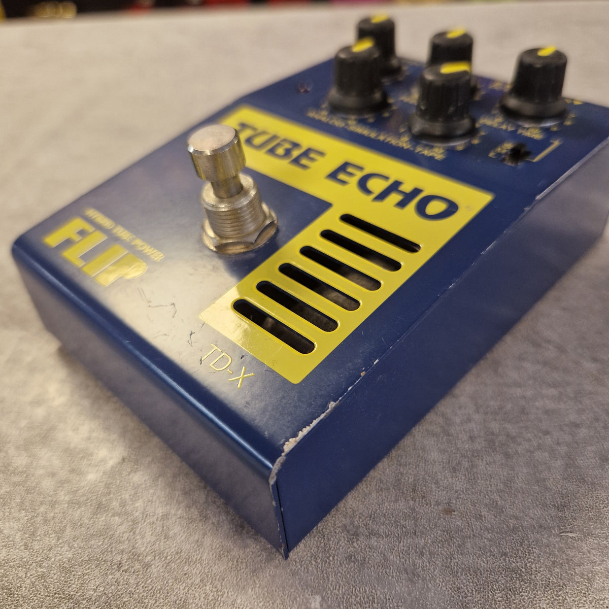 Pre-Owned Guyatone Flip TD-X Tube Echo Hybrid Tube Power Delay Pedal - A blue and yellow FXR Tube Echo pedal with black knobs and a silver footswitch, photographed on a gray textured surface.