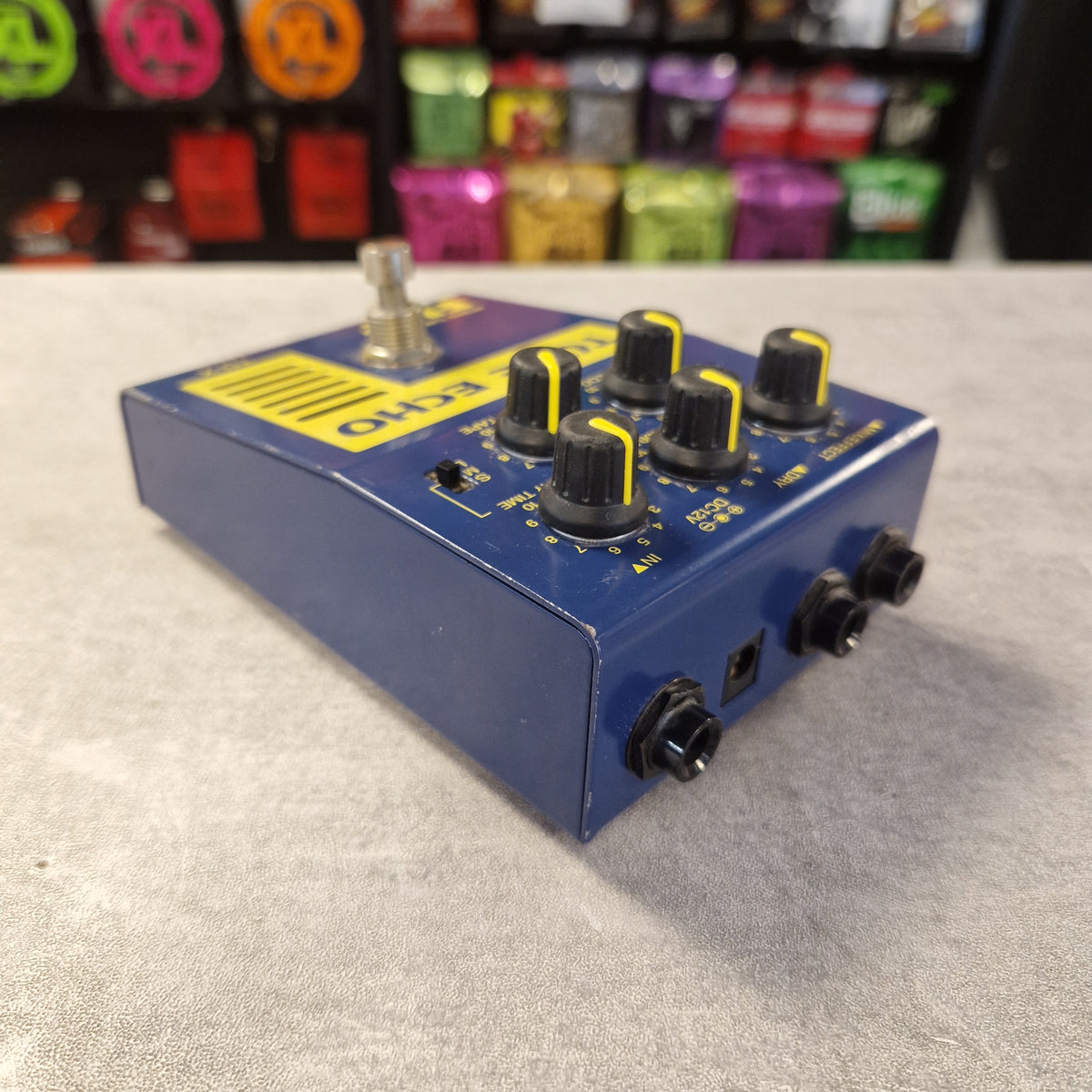 Pre-Owned Guyatone Flip TD-X Tube Echo Hybrid Tube Power Delay Pedal - A blue guitar effects pedal with black knobs and yellow accents, photographed on a light gray textured surface with a blurred store display in the background.