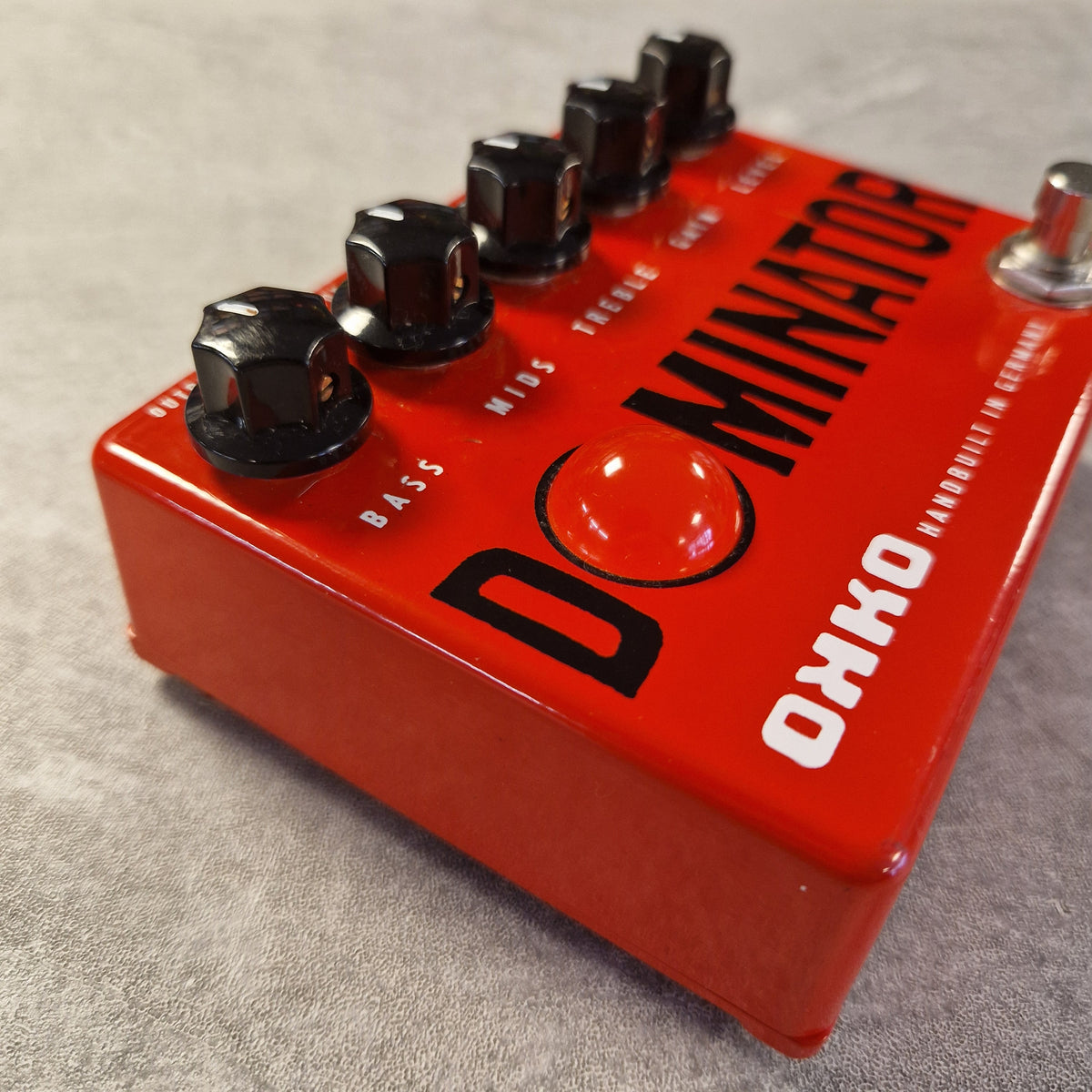 Pre-Owned Okko Dominator High Gain Distortion Pedal - A close-up of an Okko Dominator guitar effects pedal with a bright red metal body, black knobs, and white text, resting on a gray textured surface.