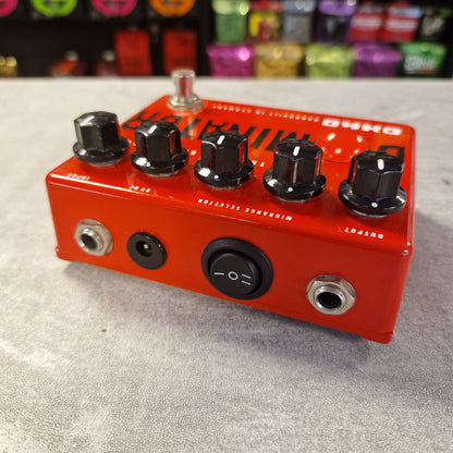 Pre-Owned Okko Dominator High Gain Distortion Pedal - A red guitar effects pedal with black knobs, silver jacks, and a silver footswitch, photographed on a gray textured surface with a blurred store background.