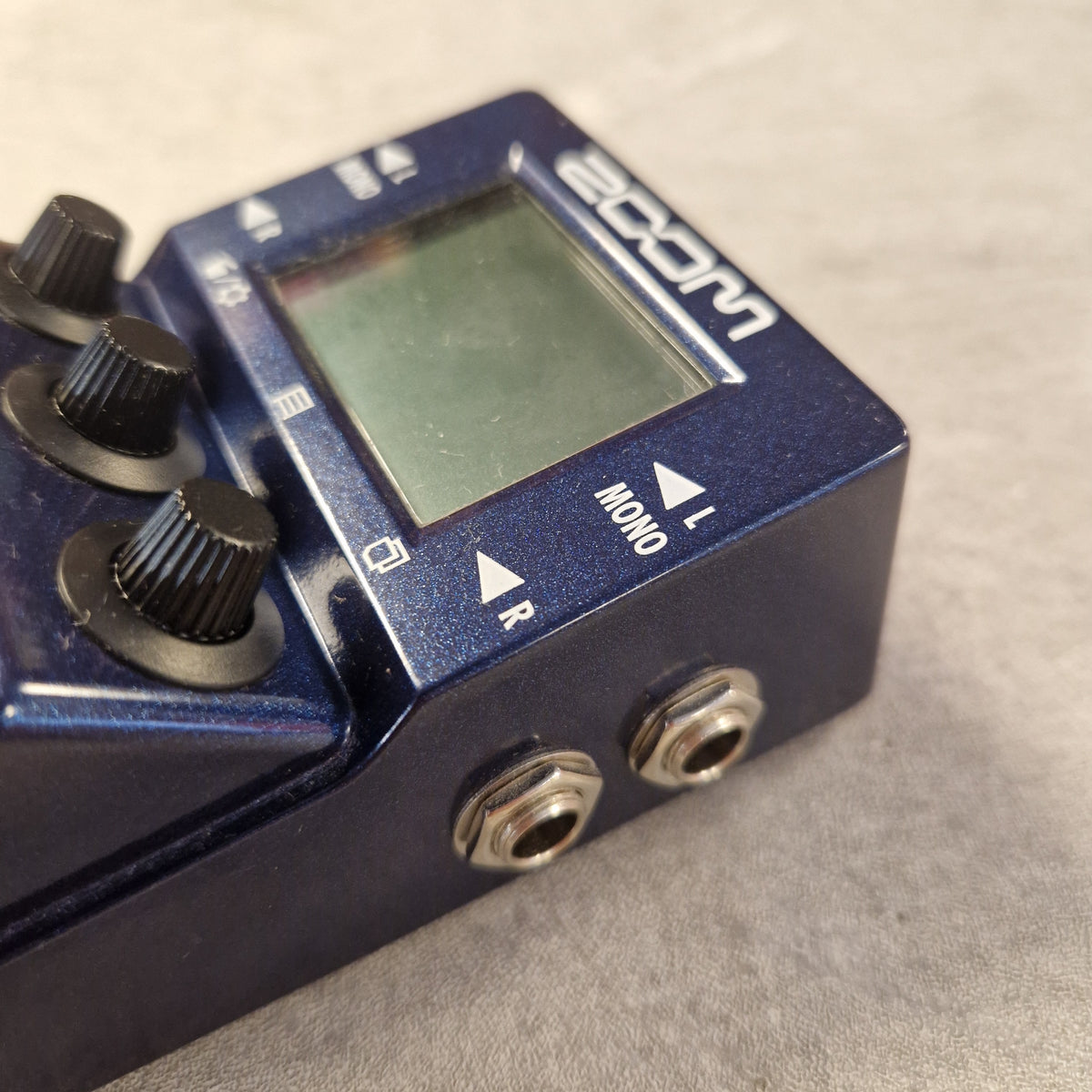 Pre-Owned Zoom MS-100BT MultiStomp Guitar Pedal with Bluetooth - A close-up shot of a blue Zoom guitar effects pedal, showing its front face with an LCD screen, three black control knobs, and two silver metal jack inputs on a textured gray background.