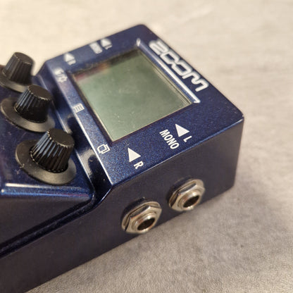 Pre-Owned Zoom MS-100BT MultiStomp Guitar Pedal with Bluetooth - A close-up shot of a blue Zoom guitar effects pedal, showing its front face with an LCD screen, three black control knobs, and two silver metal jack inputs on a textured gray background.