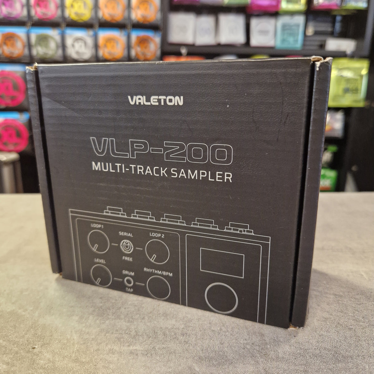 Pre-Owned Valeton VLP-200 Multi-Track Sampler w/ Box - A black Valeton VLP-200 multi-track sampler retail box standing on a gray tabletop, with a blurred store background showing shelves of colorful items.
