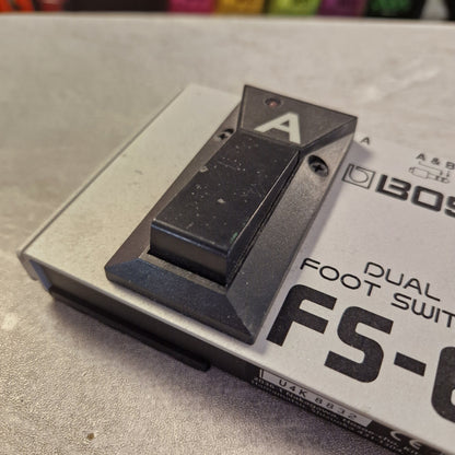 Pre-Owned Boss Dual Foot Switch FS-6 Pedal A close-up of a black dual foot switch component mounted on a silver metal base, with printed text including 'DUAL FOOT SWITCH' and partial model lettering.