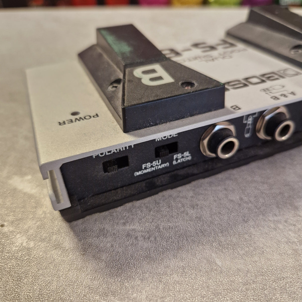 Pre-Owned Boss Dual Foot Switch FS-6 Pedal A close-up of a Boss FX-5S effects pedal showing the side panel with power switch, mode switches, and input/output jacks, resting on a gray textured surface.
