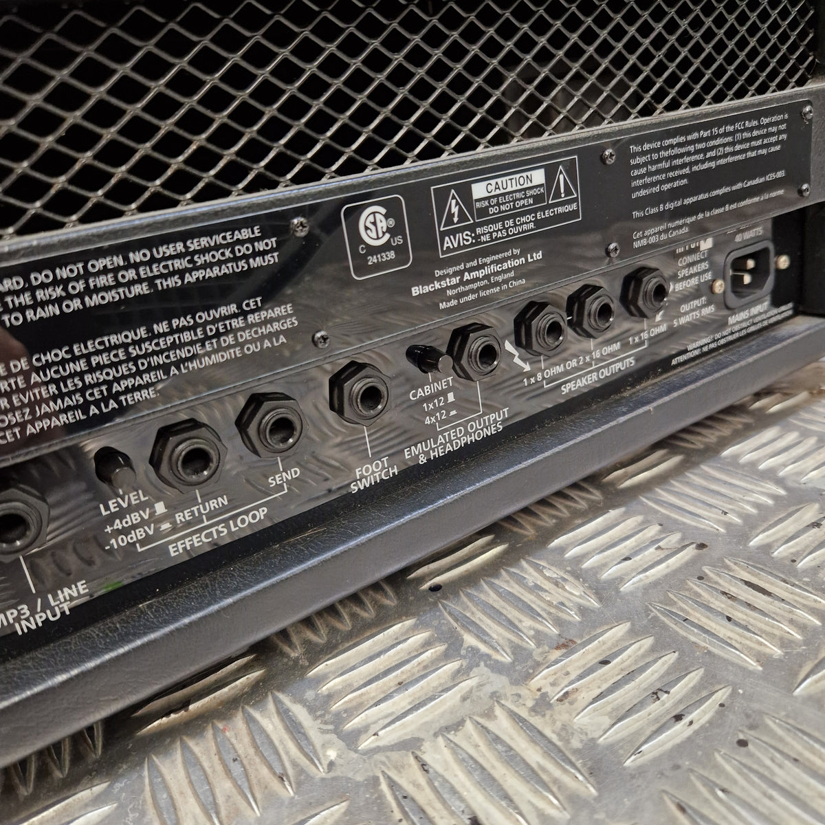 Pre-Owned Blackstar HT-5RH 5w Electric Amplifier Head w/ Footswitch - Close-up of the rear panel of a black amplifier or speaker, showing input jacks, volume controls, and labels such as 'INPUT LOOP' and 'MASTER VOL'. The panel is photographed on a metal diamond plate surface next to a mesh grille.
