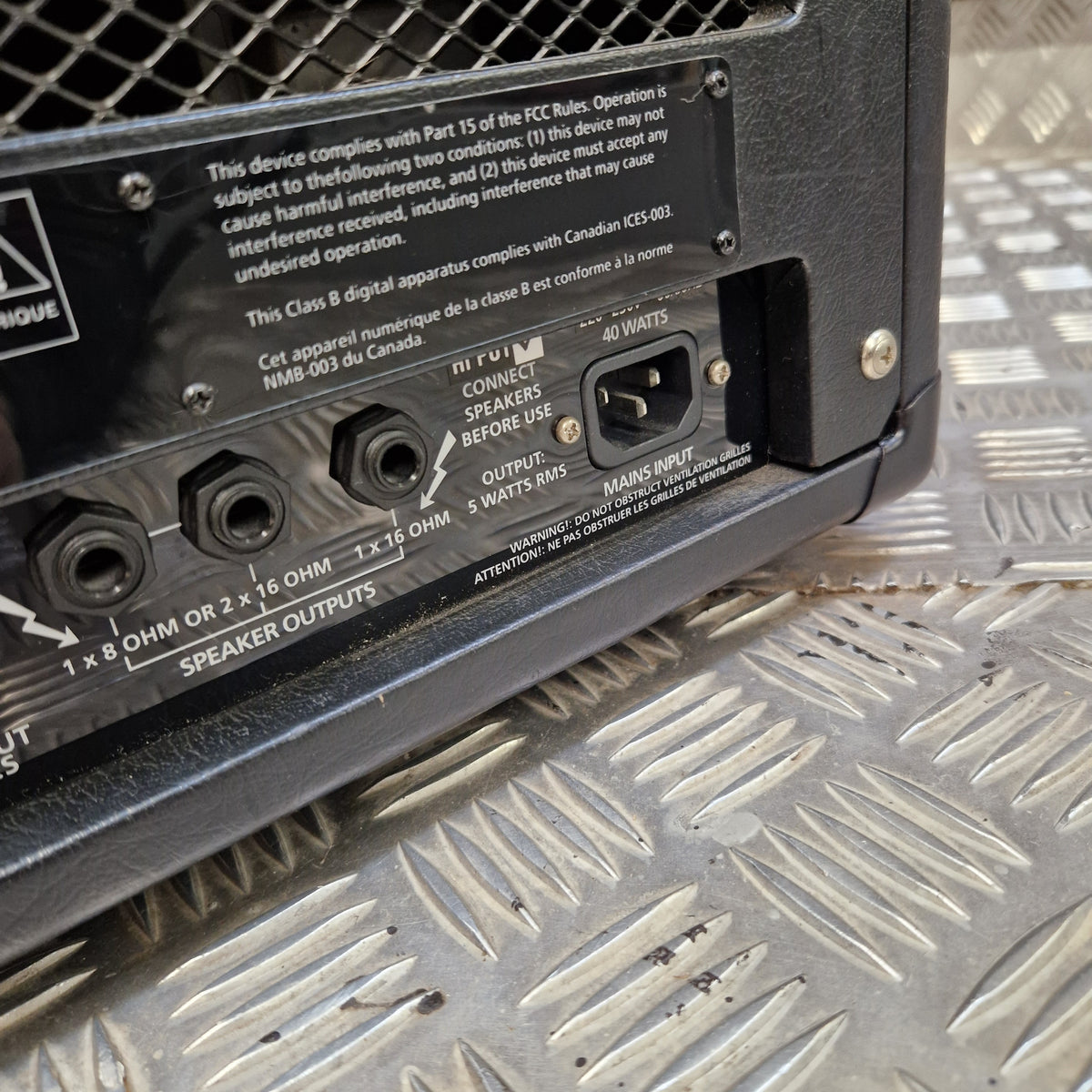 Pre-Owned Blackstar HT-5RH 5w Electric Amplifier Head w/ Footswitch - Close-up of the rear panel of a black electronic device, showing input/output jacks, power outlet, and printed technical labels, placed on a diamond plate metal surface.