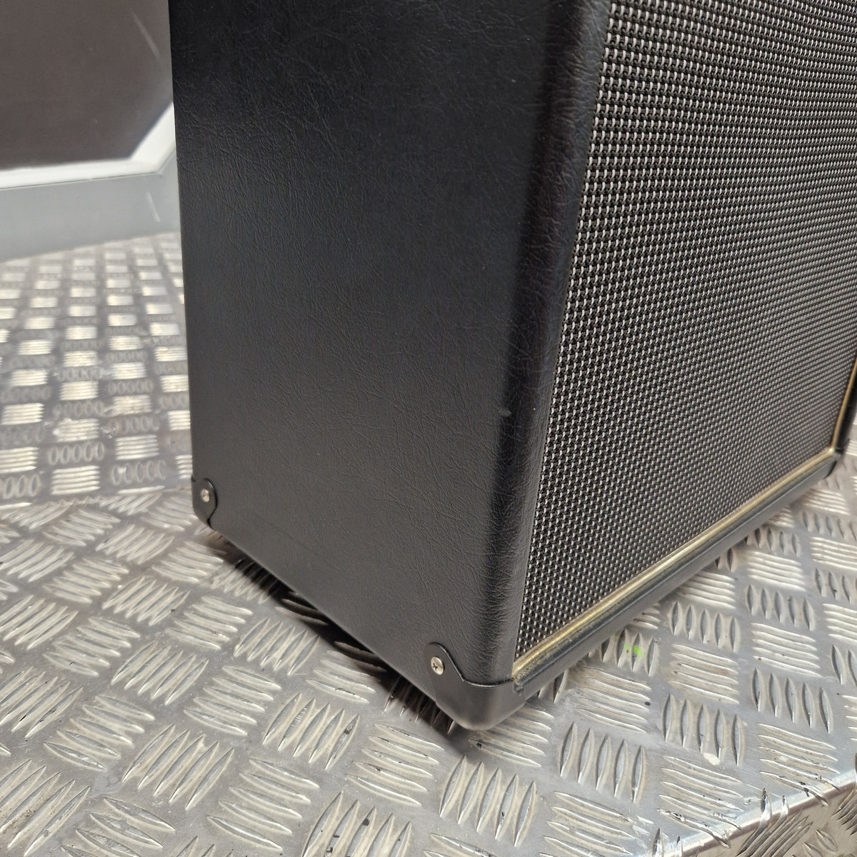 Pre-Owned Blackstar HT-112 1x12 Electric Amplifier Cab - Close-up of a black speaker cabinet corner with a metal mesh grille, photographed on a diamond-plate metal surface.