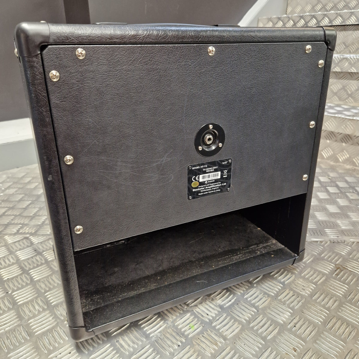 Pre-Owned Blackstar HT-112 1x12 Electric Amplifier Cab - A black textured leather or faux-leather guitar speaker cabinet shown from the rear, featuring a round binding post, a small label, and a lower speaker grille opening, photographed on a diamond plate metal floor.