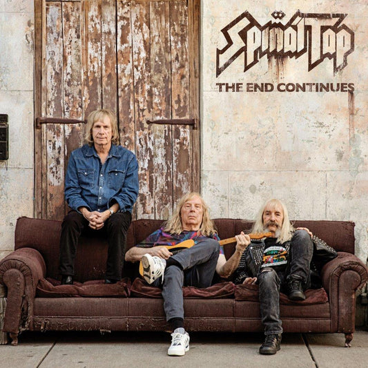 Spinal Tap - The End Continues Three musicians sitting on a couch with a rustic door and 'Spinal Tap' album cover in the background.