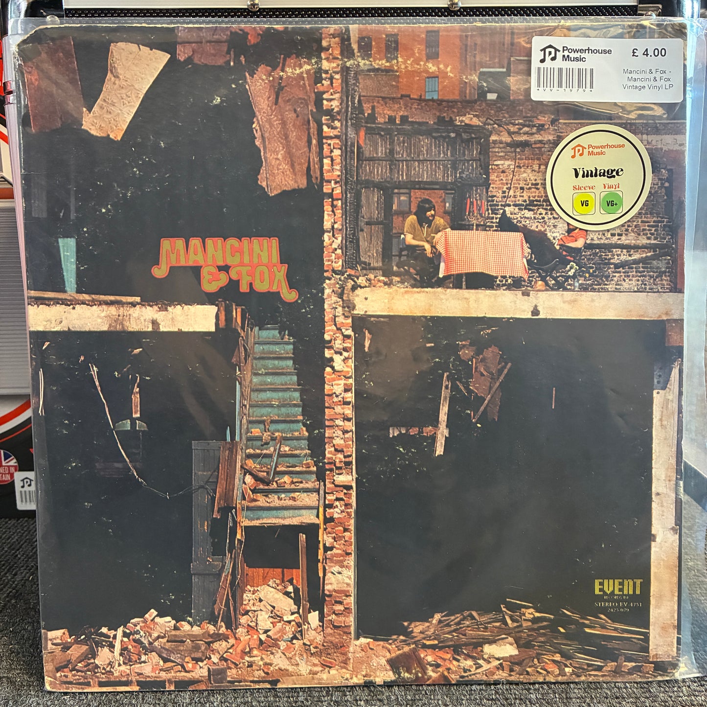 Mancini & Fox - Mancini & Fox Vintage Vinyl LP A vintage vinyl record sleeve for Marcion & Tom's 'Event' album, sealed in a protective plastic sleeve. The cover art features a dark, gritty collage of brick buildings, a staircase, and debris, with the band's name in orange text.