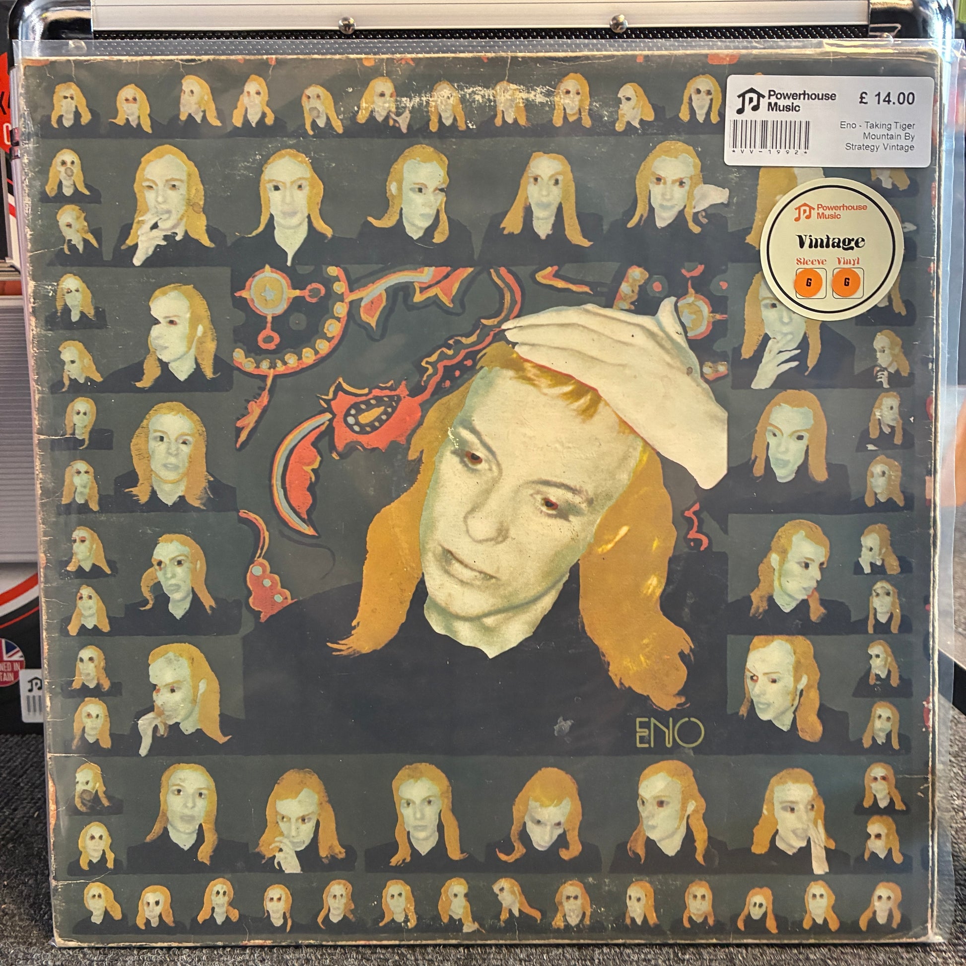 Eno - Taking Tiger Mountain By Strategy Vintage Vinyl LP A vinyl record sleeve for Enyo's 'Vintage' release, featuring a stylized portrait of a woman with orange hair against a dark background filled with smaller repeated versions of the same face. The sleeve is photographed inside a clear plastic protective case, with a barcode sticker and a circular 'Vintage' label visible on the top right.
