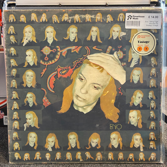 Eno - Taking Tiger Mountain By Strategy Vintage Vinyl LP A vinyl record sleeve for Enyo's 'Vintage' release, featuring a stylized portrait of a woman with orange hair against a dark background filled with smaller repeated versions of the same face. The sleeve is photographed inside a clear plastic protective case, with a barcode sticker and a circular 'Vintage' label visible on the top right.