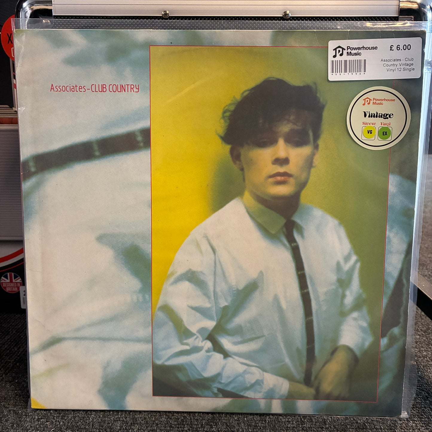Associates - Club Country Vintage Vinyl 12" Single A vinyl record sleeve for Associates-Club Country on a clear plastic shrink wrap, photographed in a record store setting. The cover features a portrait of a man in a white shirt and dark tie against a yellow background, with a pale abstract design on the left side.