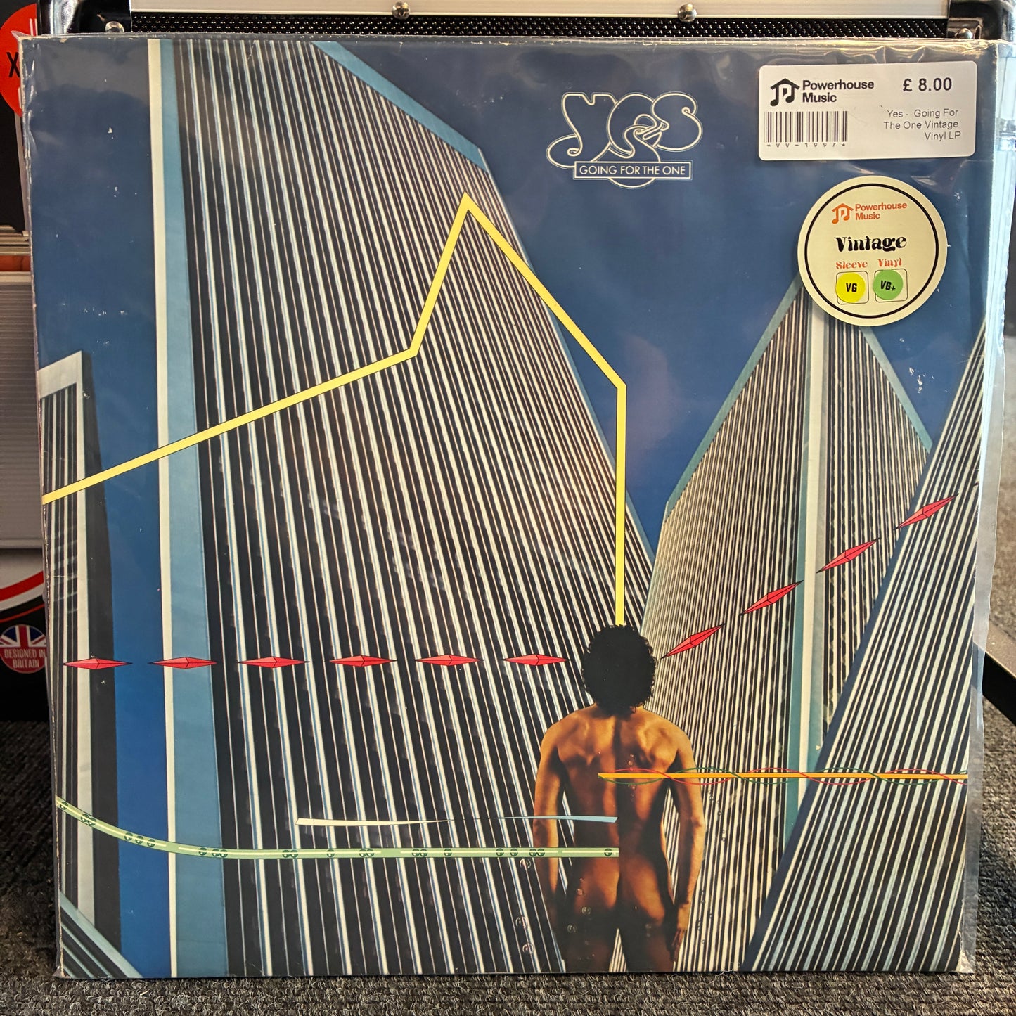 Yes -  Going For The One Vintage Vinyl LP A vinyl record sleeve for Yes's 'The Song Remembers Me' on Y2label, featuring a stylized blue cityscape with a nude figure and geometric yellow, red, and green lines. A white barcode sticker and Y2label logo are visible on the cover.