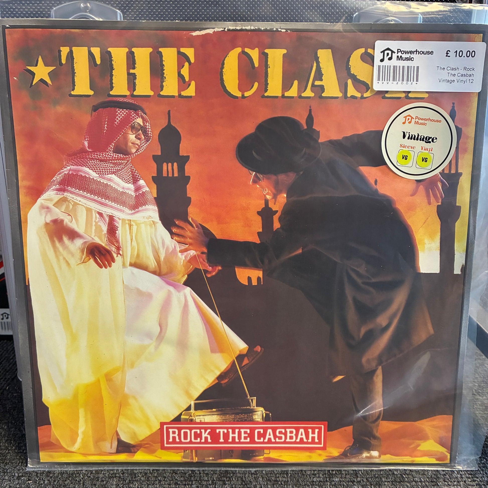 The Clash - Rock The Casbah Vintage Vinyl 12" Single A vinyl record sleeve for the band 'The Class' and album 'Rock The Casbah', showing an illustrated scene of two men in traditional Middle Eastern clothing against a warm orange sunset background. A JD Powerhouse Music 'Vintage' sticker is visible in the top right corner.