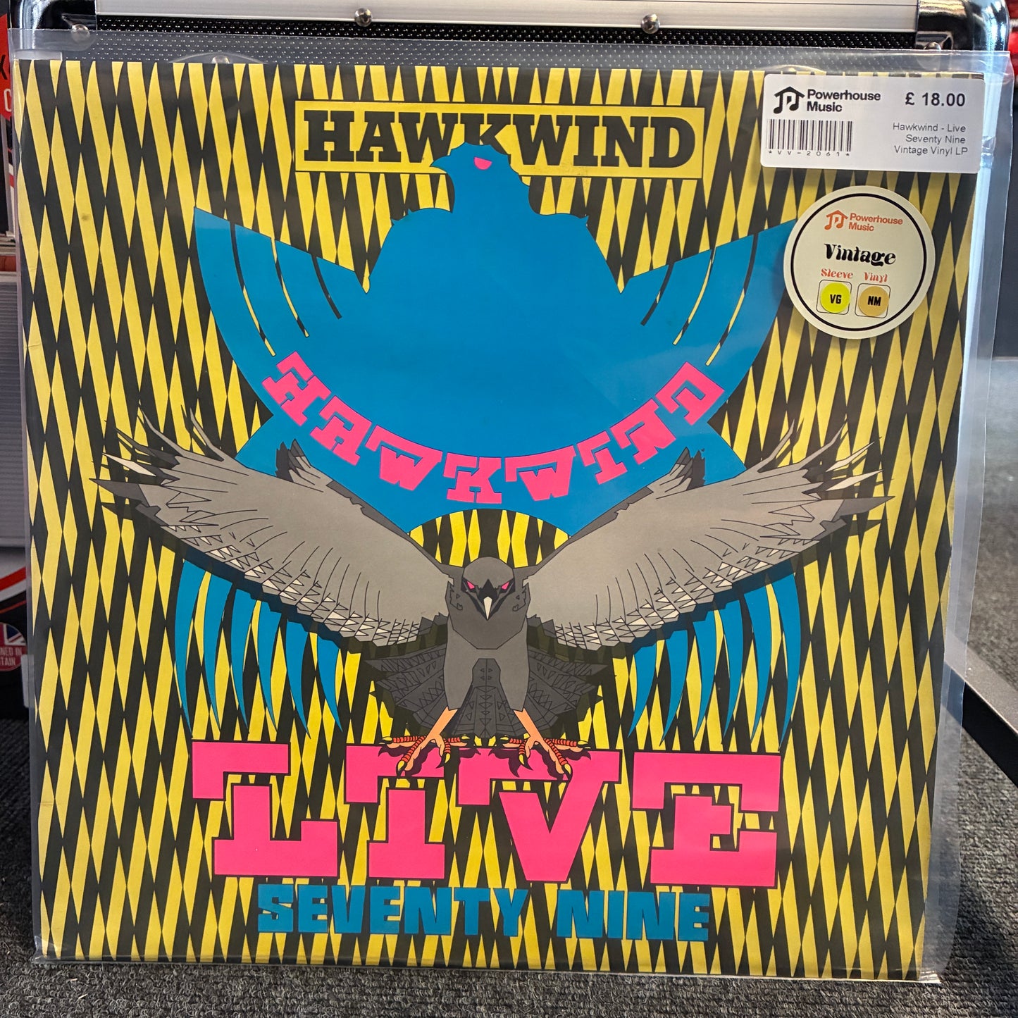 Hawkwind - Live Seventy Nine Vintage Vinyl LP A vinyl record sleeve for Hawkwind's 'Live Seventy Nine' is shown in a clear plastic sleeve, likely in a store setting. The cover art features a bold black and yellow zigzag pattern, a large blue bird silhouette, a smaller gray bird in the foreground, and pink text reading 'HAWKWind LIVE Seventy Nine'.