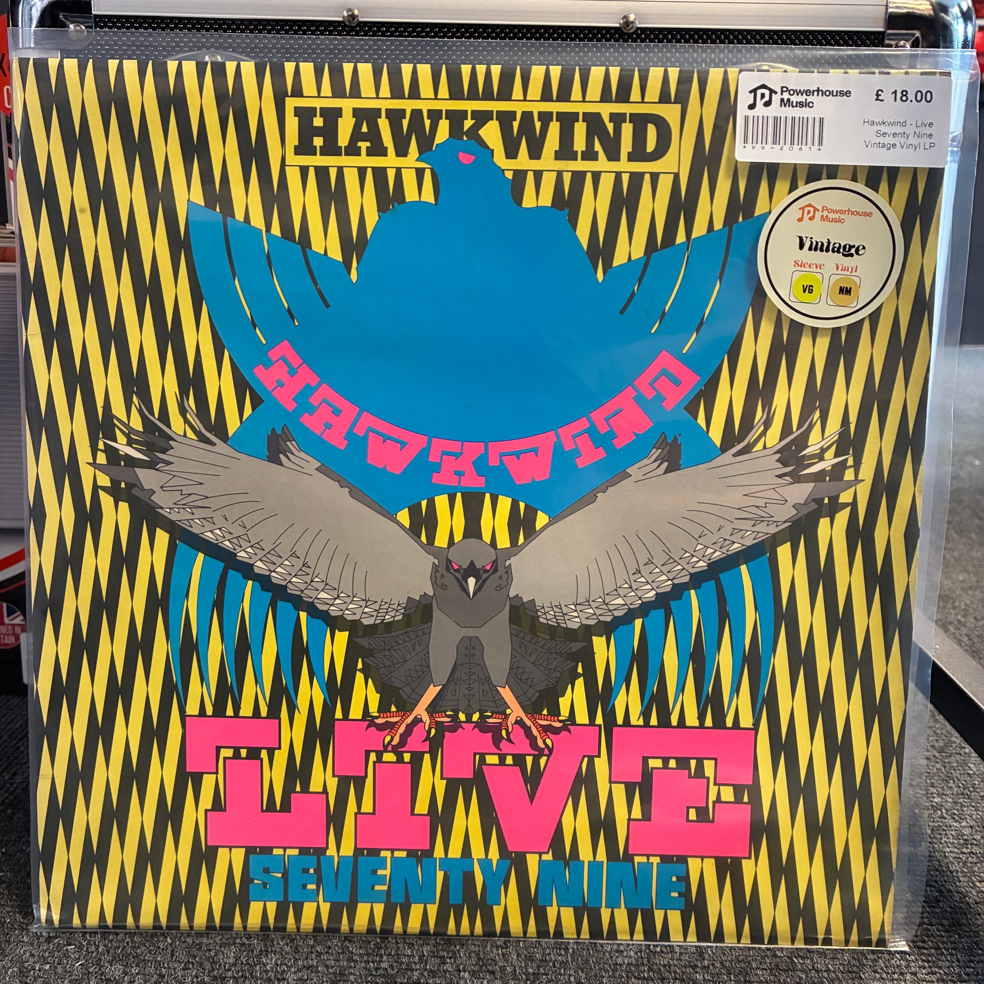 Hawkwind - Live Seventy Nine Vintage Vinyl LP A vinyl record sleeve for Hawkwind's 'Live Seventy Nine' is shown in a clear plastic sleeve, likely in a store setting. The cover art features a bold black and yellow zigzag pattern, a large blue bird silhouette, a smaller gray bird in the foreground, and pink text reading 'HAWKWind LIVE Seventy Nine'.
