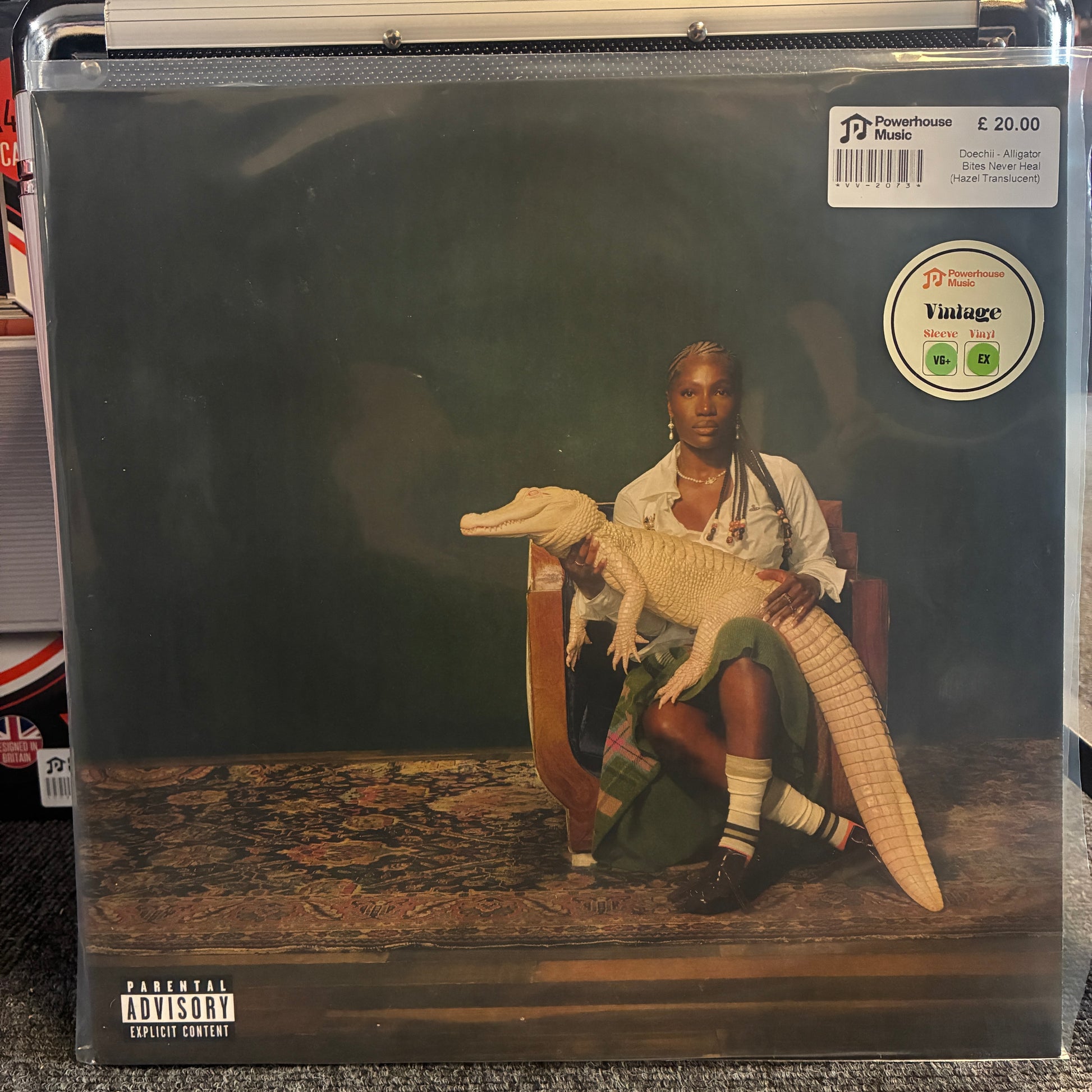 Doechii - Alligator Bites Never Heal (Hazel Translucent) Vintage Vinyl A vinyl record sleeve for Kendrick Lamar's 'The King and the Queen' is displayed in a clear protective plastic sleeve, likely from a record store. The cover art features a man in a white shirt and green skirt sitting in a chair with a large alligator.
