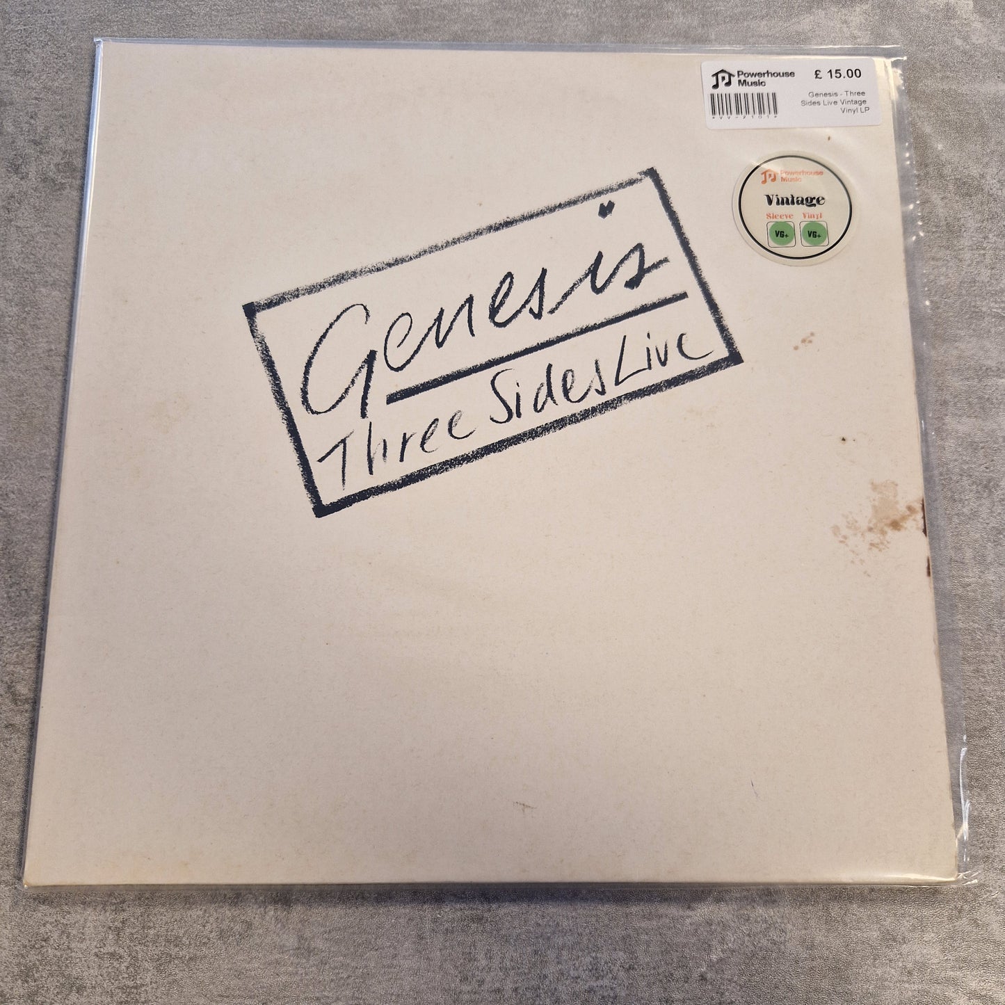 Genesis - Three Sides Live Vintage Vinyl LP A square album cover for the band Genesis, featuring the text 'Three Side Lines' in black script inside a hand-drawn border. The cover is off-white with a visible scuff and a barcode sticker on the top left, photographed on a gray textured surface.