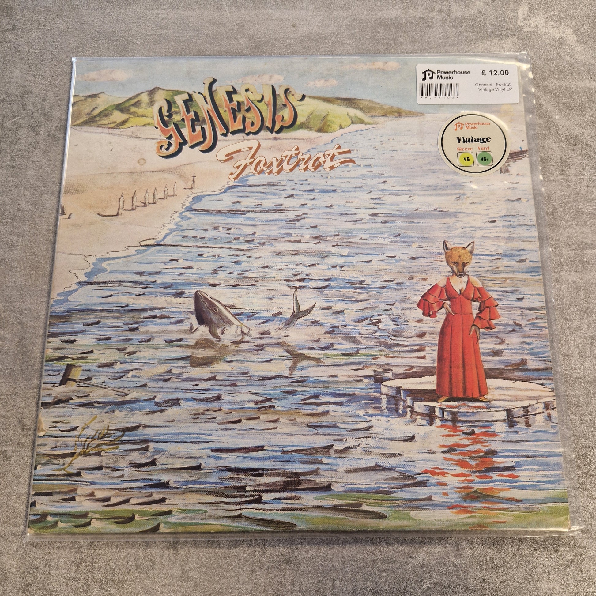 Genesis - Foxtrot Vintage Vinyl LP A square printed item wrapped in clear plastic, featuring an illustrated coastal scene with a figure in a red dress standing on a pier and several dolphins in the water. The left side has stylized vertical text, and a barcode sticker is visible in the upper left.