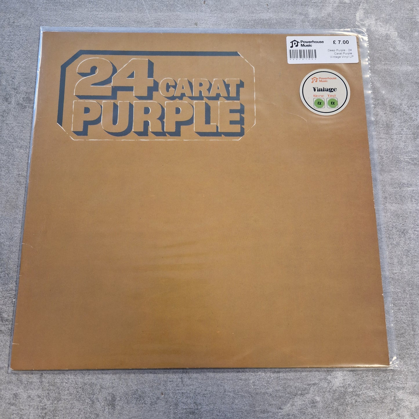 Deep Purple - 24 Carat Purple Vintage Vinyl LP A 24 Carat Purple vinyl record sleeve in its clear plastic shrink wrap, placed on a gray textured surface. The sleeve is a metallic gold color with the band's name printed in a stylized dark blue font.