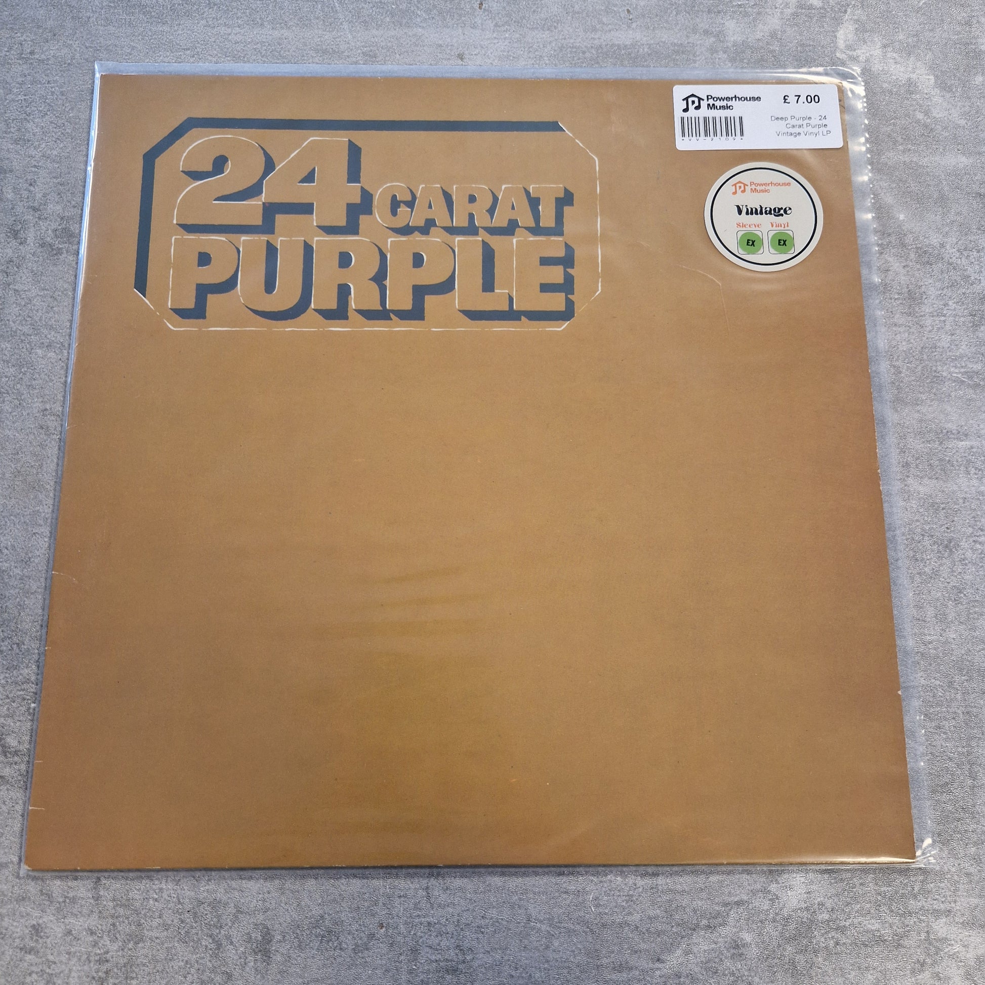 Deep Purple - 24 Carat Purple Vintage Vinyl LP A 24 Carat Purple vinyl record sleeve in its clear plastic shrink wrap, placed on a gray textured surface. The sleeve is a metallic gold color with the band's name printed in a stylized dark blue font.