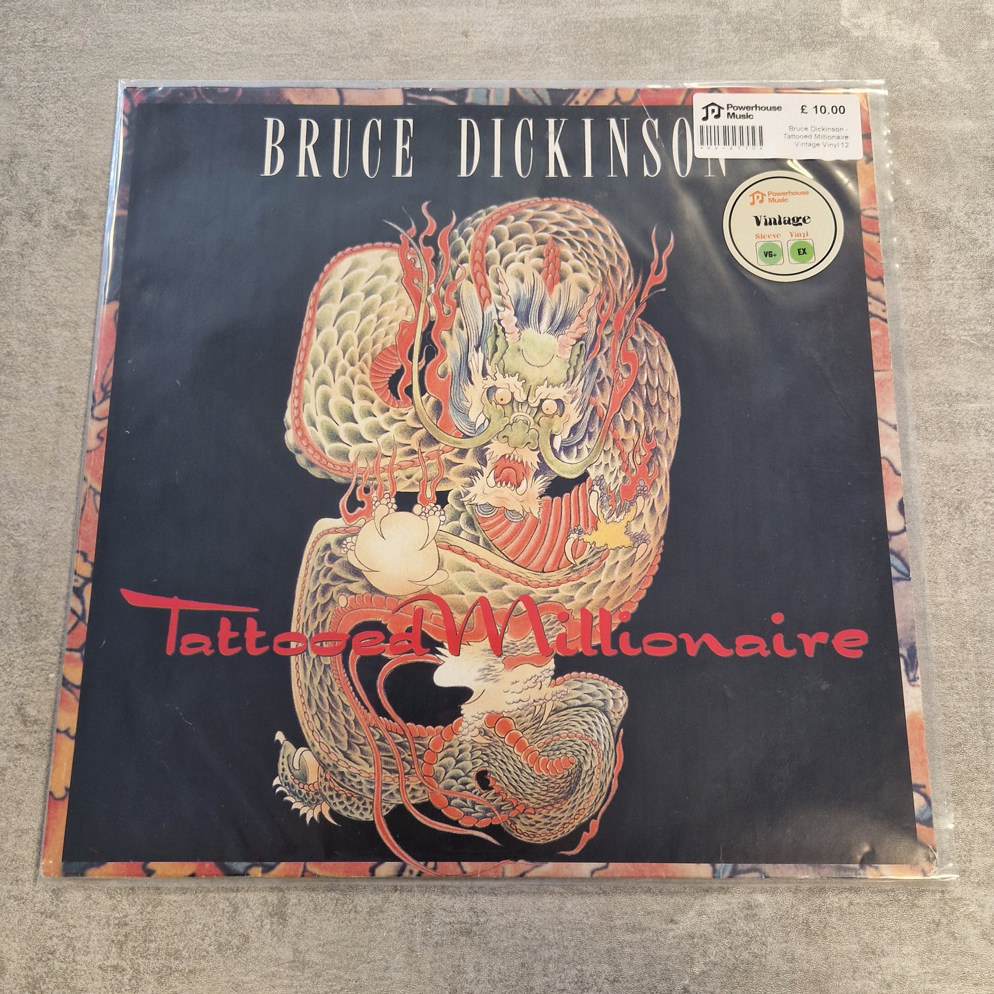 Bruce Dickinson - Tattooed Millionaire Vintage Vinyl 12" Single A vinyl record sleeve for Bruce Dickinson's 'Tattooed Millionaire' album, featuring a detailed red and gold dragon illustration on a black background, photographed on a gray textured surface.
