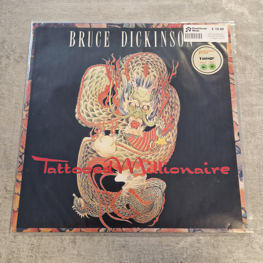 Bruce Dickinson - Tattooed Millionaire Vintage Vinyl 12" Single A vinyl record sleeve for Bruce Dickinson's 'Tattooed Millionaire' album, featuring a detailed red and gold dragon illustration on a black background, photographed on a gray textured surface.