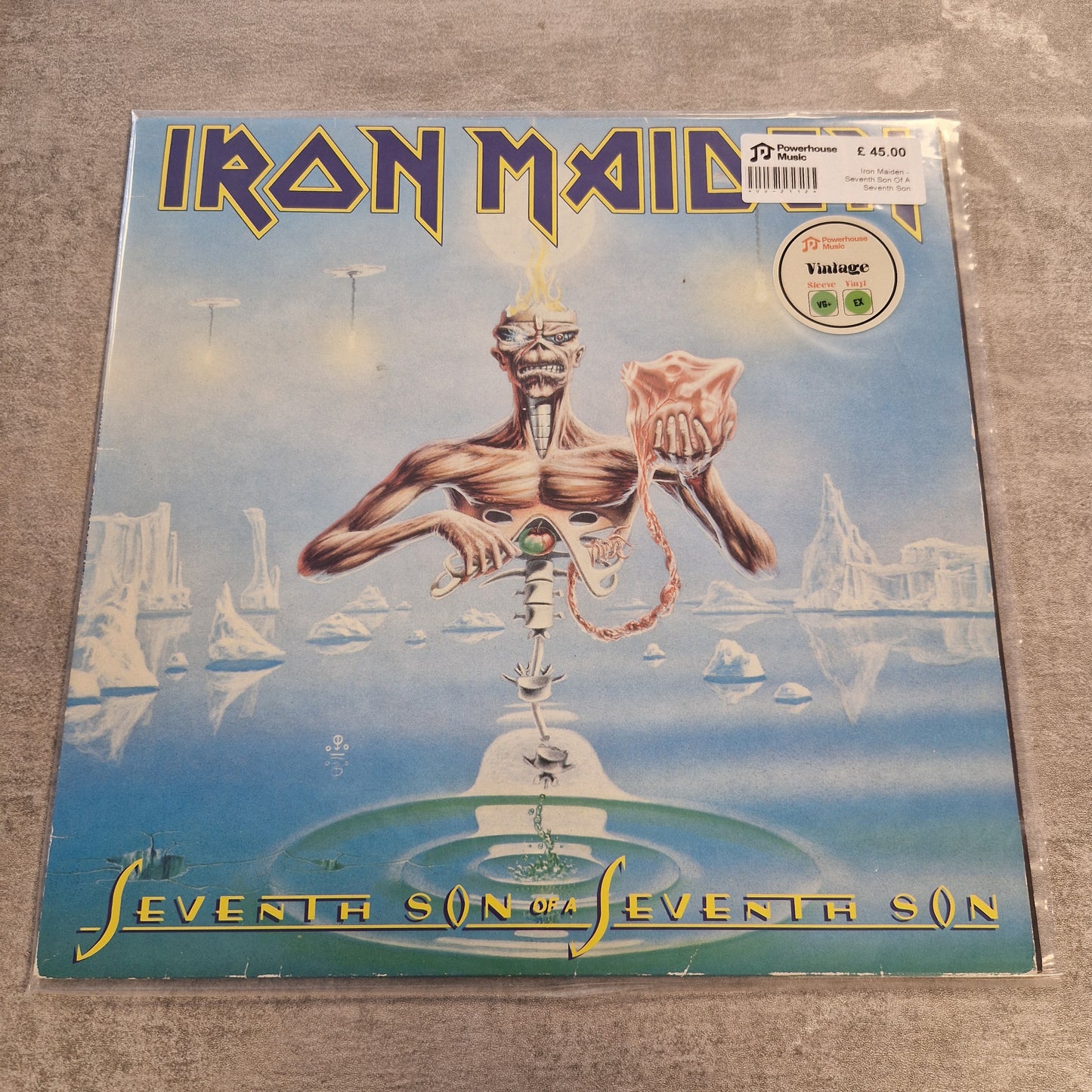 Iron Maiden - Seventh Son Of A Seventh Son Vintage Vinyl LP Iron Maiden 'Seveneth Son' vinyl record sleeve photographed on a textured gray surface. The cover art shows a surreal blue landscape with a floating humanoid figure and the band name 'Iron Maiden' in large gold lettering on the left.