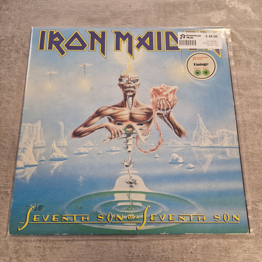 Iron Maiden - Seventh Son Of A Seventh Son Vintage Vinyl LP Iron Maiden 'Seveneth Son' vinyl record sleeve photographed on a textured gray surface. The cover art shows a surreal blue landscape with a floating humanoid figure and the band name 'Iron Maiden' in large gold lettering on the left.
