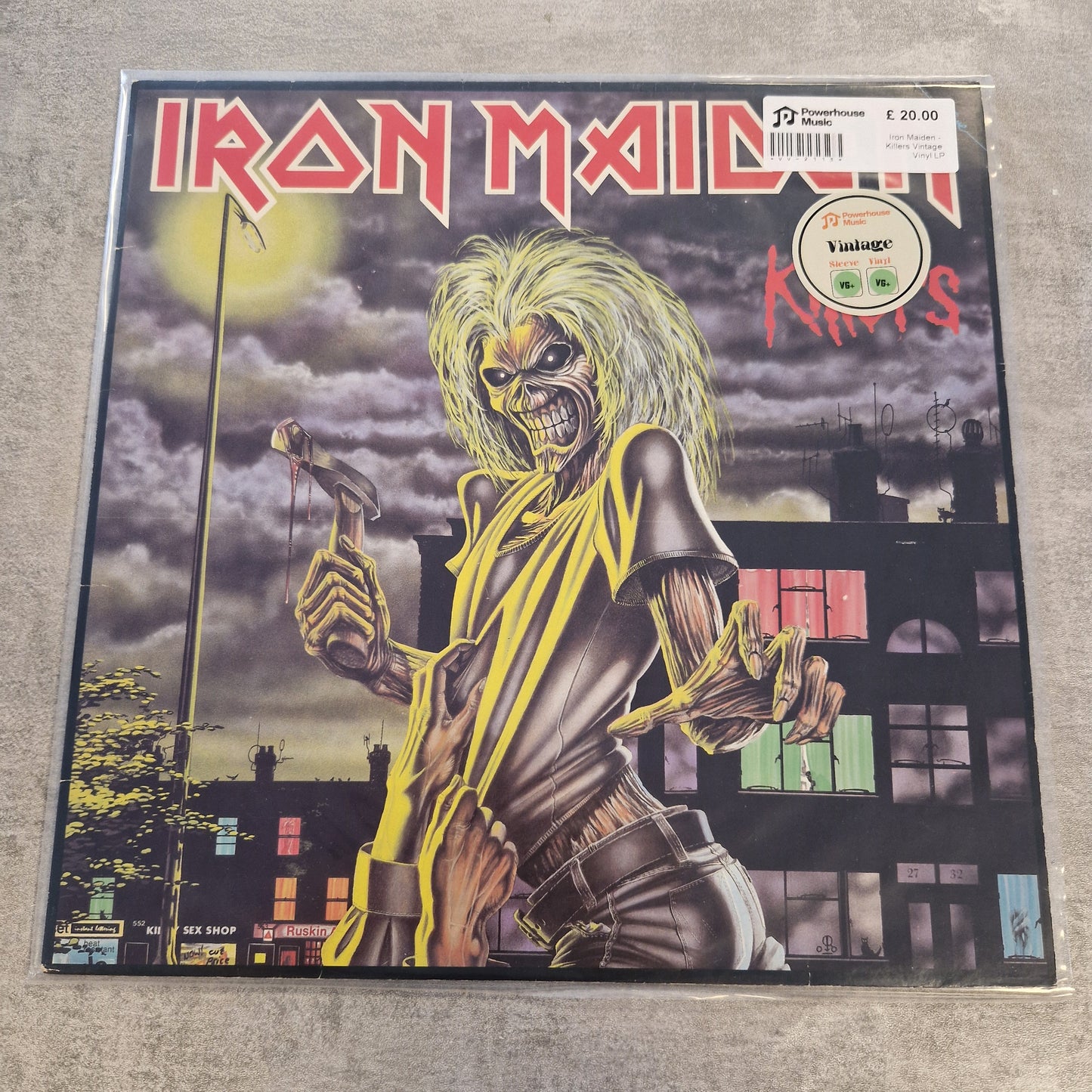 Iron Maiden - Killers Vintage Vinyl LP Iron Maiden 'Miles' vinyl record sleeve on a clear protective plastic case, photographed flat on a gray textured surface. The cover art features the band's mascot, Eddie, with wild yellow hair and a cymbal against a dark cityscape background.