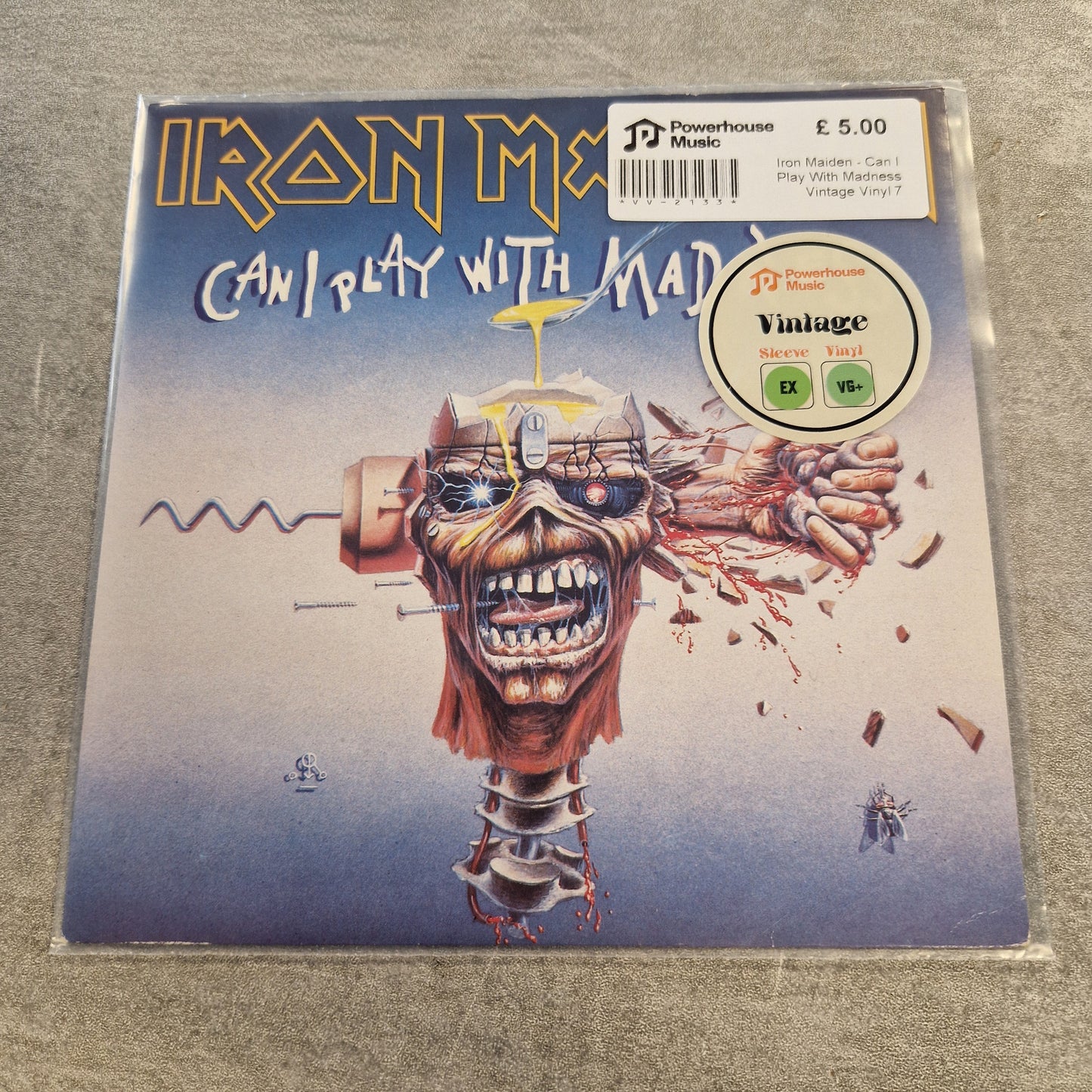 Iron Maiden - Can I Play With Madness Vintage Vinyl 7" Single Iron Maiden 'Can't Play With a Mad' vinyl record sleeve in a clear plastic sleeve, photographed on a gray textured surface. The cover art features a surreal, exploded portrait of the mascot Eddie with a brain and mechanical details, with the band's name and album title in large stylized text.