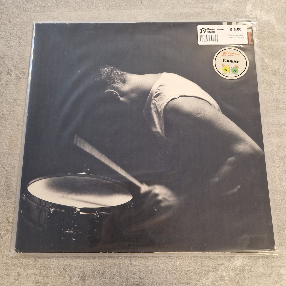 U2 - Desire Vintage Vinyl 12" Single A vinyl record sleeve for Django Reinhardt's 'Vintages' LP, featuring a dramatic black and white photograph of the drummer playing. The record is sealed in plastic and sits on a gray concrete floor, with a white 'Vintages' sticker and a barcode sticker on the top right.
