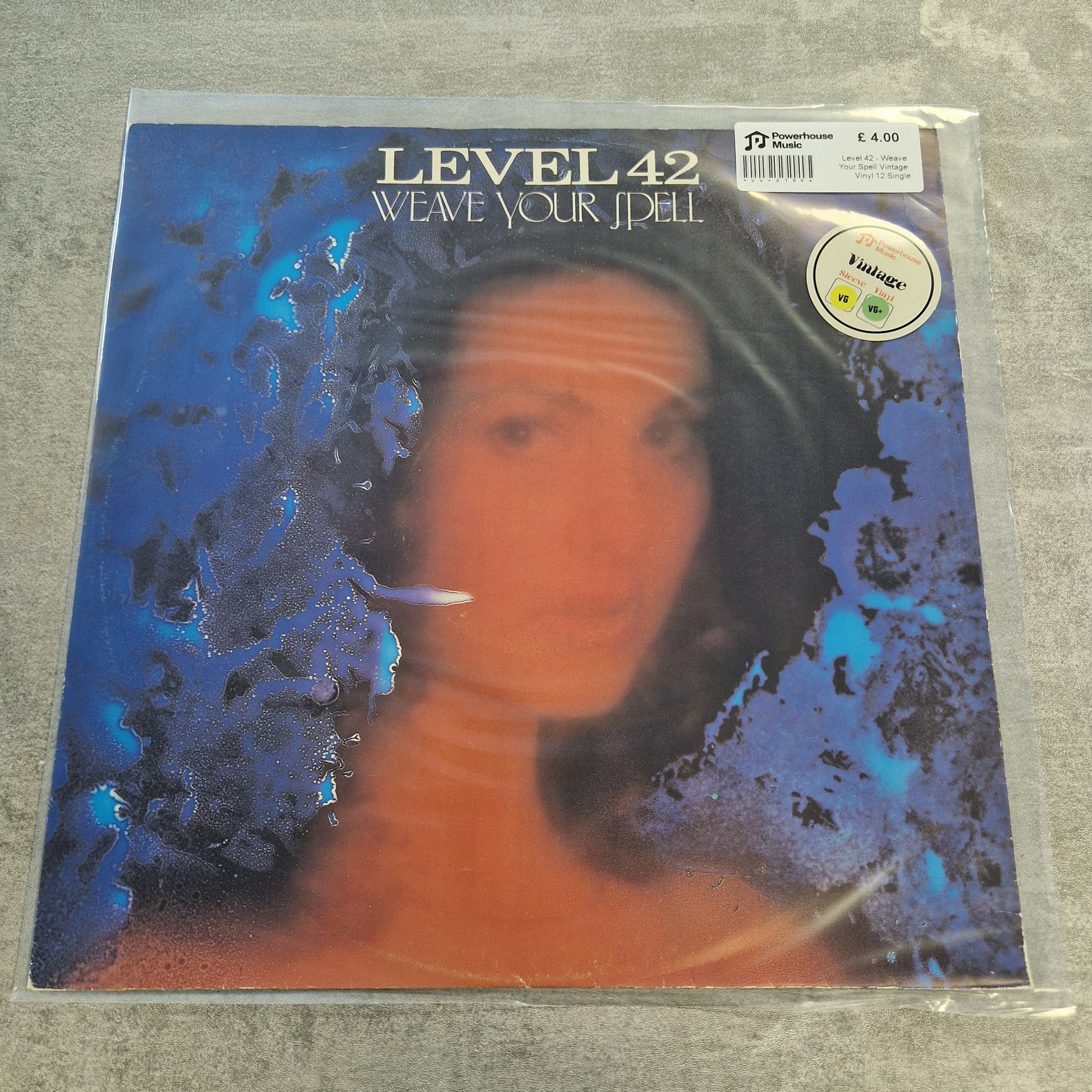 Level 42 - Weave Your Spell Vintage Vinyl 12" Single A vinyl record sleeve for Level 42's 'Weave Your Jell' is sealed in a clear plastic sleeve and photographed on a gray textured surface. The cover art features a portrait of a woman with a blue and orange color treatment, and a silver foil sticker is visible on the top right.