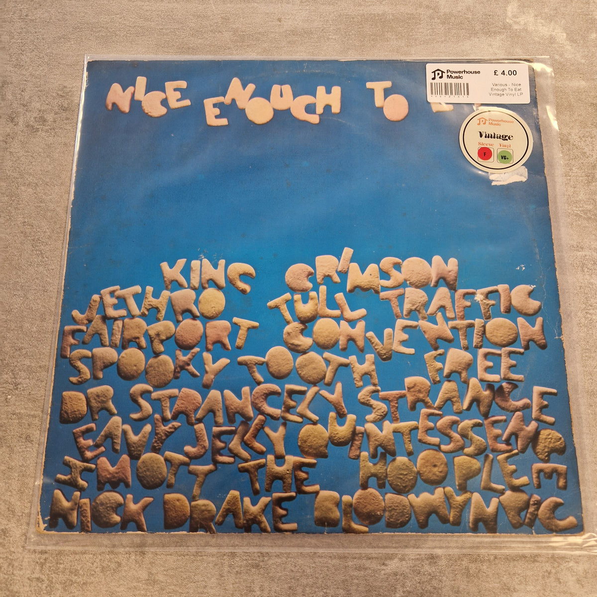 Various - Nice Enough To Eat Vintage Vinyl LP A vinyl record sleeve for the band 'Nice Enough To' featuring a bright blue background with the band's name in beige at the top and the names of various comedy clubs in raised beige lettering across the bottom. The sleeve is photographed flat on a gray textured surface, with a barcode sticker and a Yastigoo sticker visible in the top right corner.
